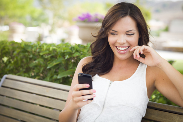 Attractive Smiling Young Adult Female Texting on Cell Phone Outdoors on a Bench.