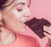 Beautiful young happy woman with a bar of chocolate on a pink background and bright makeup, a close-up frame takes a bite